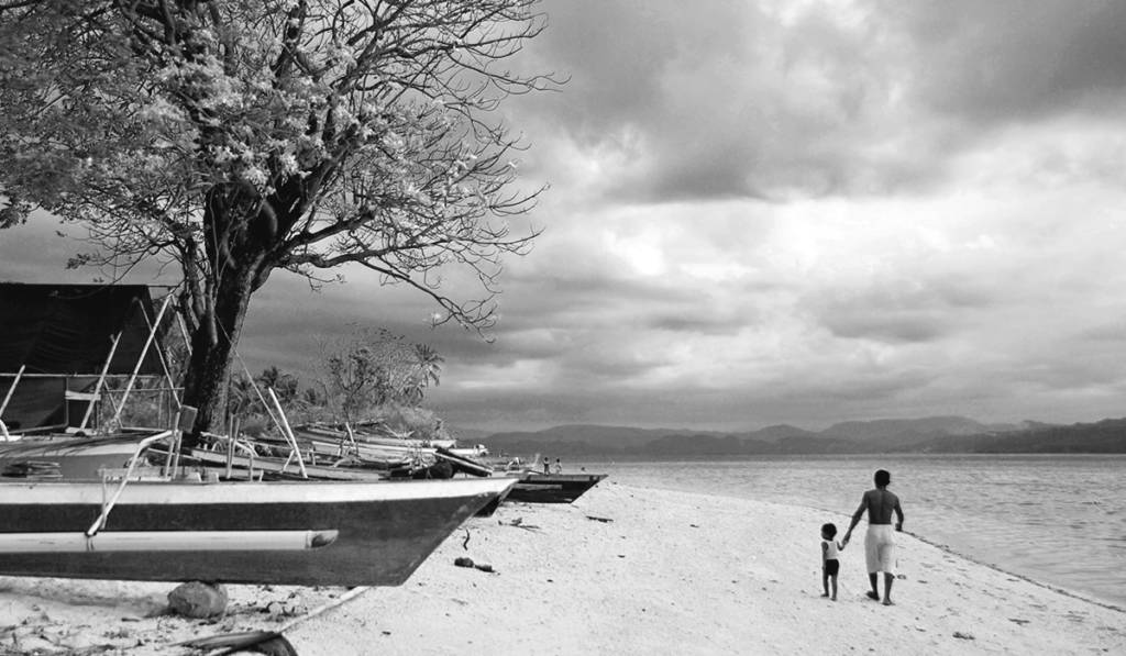 Mann und Kind spazieren am Meer, Baum und Boote am Strand