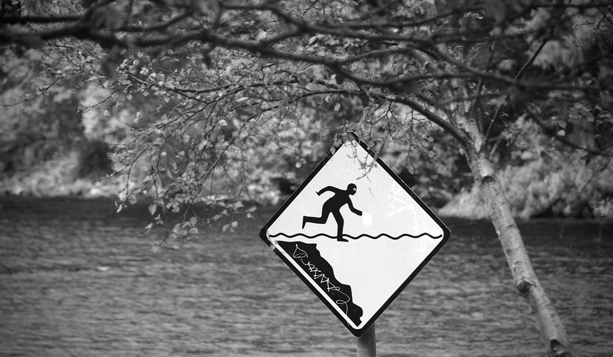 Ein Schild, das eine Person zeigt, die, wenn sie weiterläuft, ins Wasser fällt. Dahinter Wasser. Im Vordergrund ein Baum.
